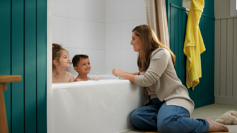 Mother smiling and interacting with two young children during bath time in a Bath Fitter white acrylic bathtub, surrounded by teal cabinetry and neutral bathroom decor
