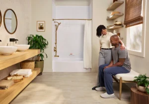 A man and woman relaxing in a bright, modern bathroom featuring a Bath Fitter glass-enclosed shower with brass hardware, cream walls, and light wood flooring